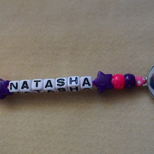 May include: A purple and pink beaded keychain with the name "Natasha" spelled out in white letter beads. A round charm with a picture of a girl with long brown hair is attached to the end of the keychain.