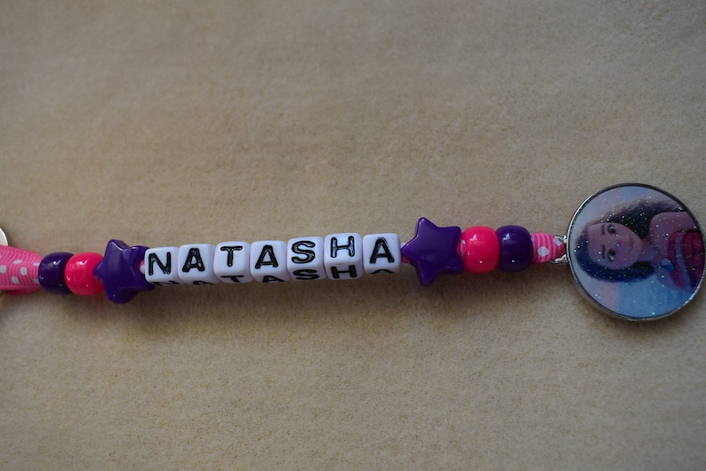 May include: A purple and pink beaded keychain with the name "Natasha" spelled out in white letter beads. A round charm with a picture of a girl with long brown hair is attached to the end of the keychain.