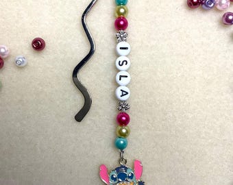 May include: A handmade bookmark featuring a silver metal hook and a beaded strand. The strand includes colorful beads, white letter beads spelling "ISLA", and a charm of a blue cartoon character. Additional beads are scattered around.