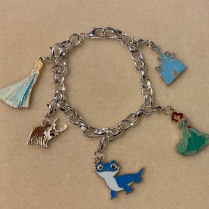 May include: Silver charm bracelet with enamel charms. The charms depict a blue castle, a blue and white dress, a brown reindeer, a blue lizard, and a green dress with red hair. The bracelet is displayed on a neutral surface.