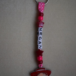 May include: A keyring featuring a silver metal ring attached to a pink ribbon with white polka dots. The strand includes red and pink beads, two red star-shaped beads, and white cube beads spelling the name MEGAN, ending with a character charm.
