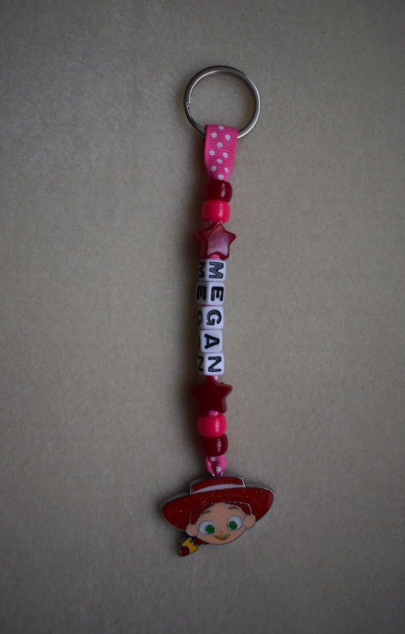 May include: A keyring featuring a silver metal ring attached to a pink ribbon with white polka dots. The strand includes red and pink beads, two red star-shaped beads, and white cube beads spelling the name MEGAN, ending with a character charm.