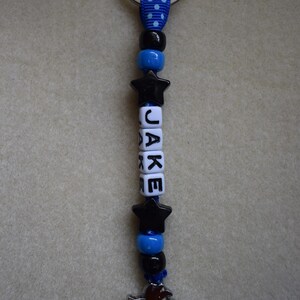 May include: A keyring featuring a silver metal ring attached to a blue ribbon with white polka dots. The strand includes black and blue beads, two black star-shaped beads, and white cube beads spelling the name JAKE, ending with a horse charm.