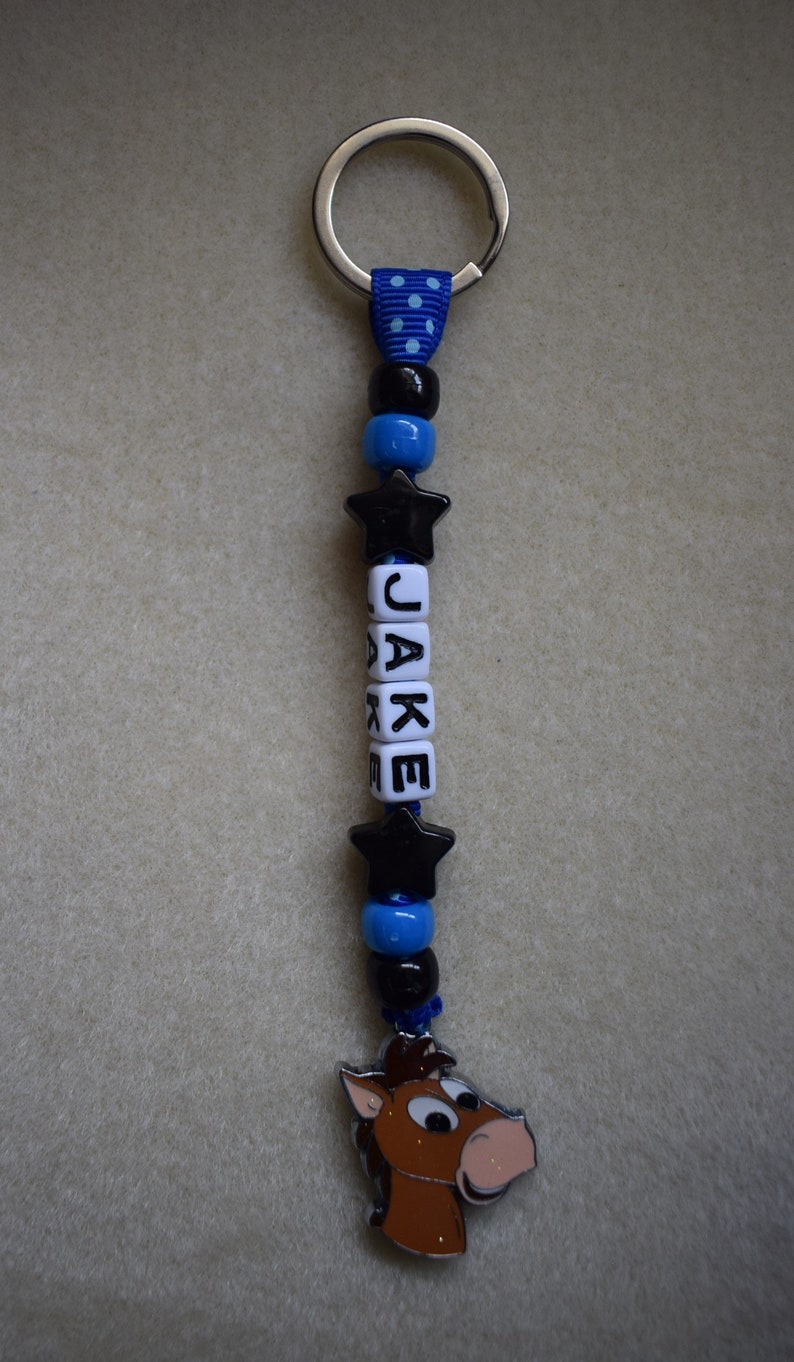 May include: A keyring featuring a silver metal ring attached to a blue ribbon with white polka dots. The strand includes black and blue beads, two black star-shaped beads, and white cube beads spelling the name JAKE, ending with a horse charm.