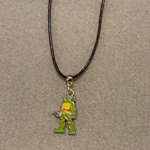 May include: A necklace featuring a dark brown cord and a silver-toned metal pendant shaped like a green turtle character. The character wears an orange mask, orange elbow and knee pads, and holds a silver-coloured weapon in its right hand.
