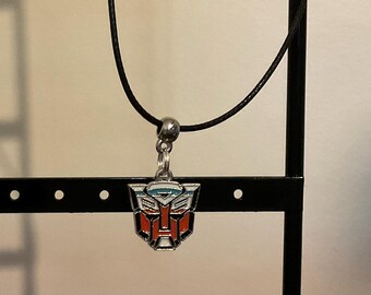 May include: A silver pendant necklace with the Autobot symbol, featuring red, white, blue, and silver enamel. The pendant is suspended from a black cord necklace with a silver bead clasp. The necklace is displayed on a black stand.