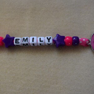 May include: A pink and purple beaded keychain with the name "EMILY" spelled out in black letters on white beads. The keychain also features a pink star charm and a pink round charm with a cartoon character.