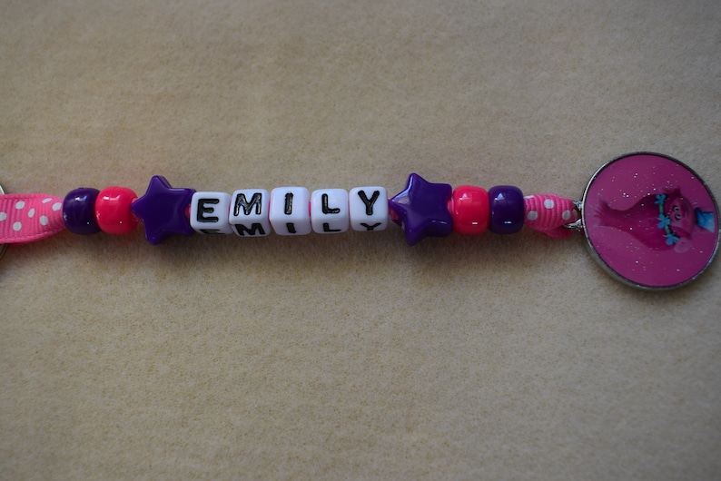 May include: A pink and purple beaded keychain with the name "EMILY" spelled out in black letters on white beads. The keychain also features a pink star charm and a pink round charm with a cartoon character.