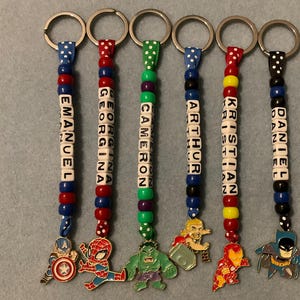 Peut inclure: Six porte-clés avec des noms écrits sur des perles. Chaque porte-clés a un code couleur différent et un charm représentant un super-héros. Les noms sont Emanuel, Georgina, Cameron, Arthur, Kristian et Daniel.