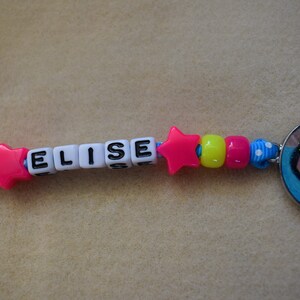 May include: A pink star charm with the name "ELISE" spelled out in black letters on white beads. The charm is attached to a blue and white ribbon with yellow and pink beads.
