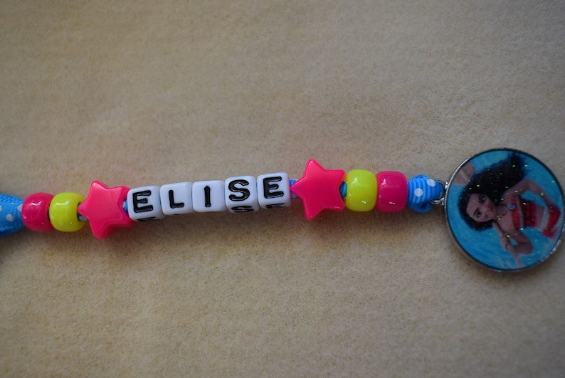 May include: A pink star charm with the name "ELISE" spelled out in black letters on white beads. The charm is attached to a blue and white ribbon with yellow and pink beads.
