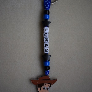 May include: A keyring featuring a silver metal ring attached to a blue polka-dot ribbon. The ribbon is strung with alternating black and blue beads, two black star-shaped beads, and white letter beads spelling LUKAS, ending in a Woody charm.