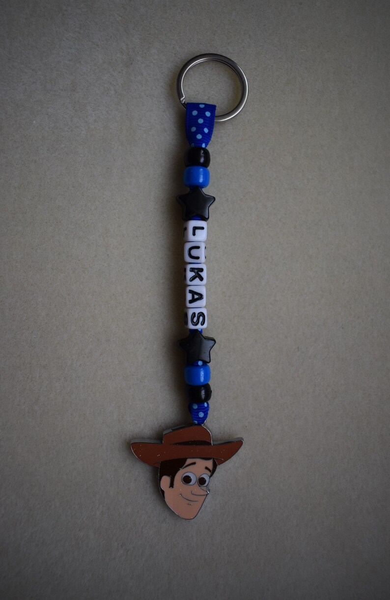 May include: A keyring featuring a silver metal ring attached to a blue polka-dot ribbon. The ribbon is strung with alternating black and blue beads, two black star-shaped beads, and white letter beads spelling LUKAS, ending in a Woody charm.