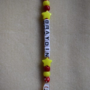 May include: A keyring featuring a silver metal ring attached to a red and white polka dot ribbon. The strand includes yellow star-shaped beads, red and yellow round beads, and white square letter beads spelling BRAYDIN, ending with a metal charm.