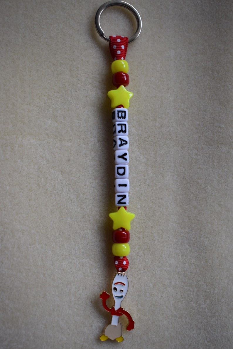 May include: A keyring featuring a silver metal ring attached to a red and white polka dot ribbon. The strand includes yellow star-shaped beads, red and yellow round beads, and white square letter beads spelling BRAYDIN, ending with a metal charm.