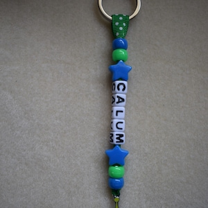 May include: A keyring featuring a green ribbon with white polka dots, blue and green plastic beads, and white cube beads spelling the name CALUM. The design includes two blue star-shaped beads and a metal charm of a three-eyed green alien character.