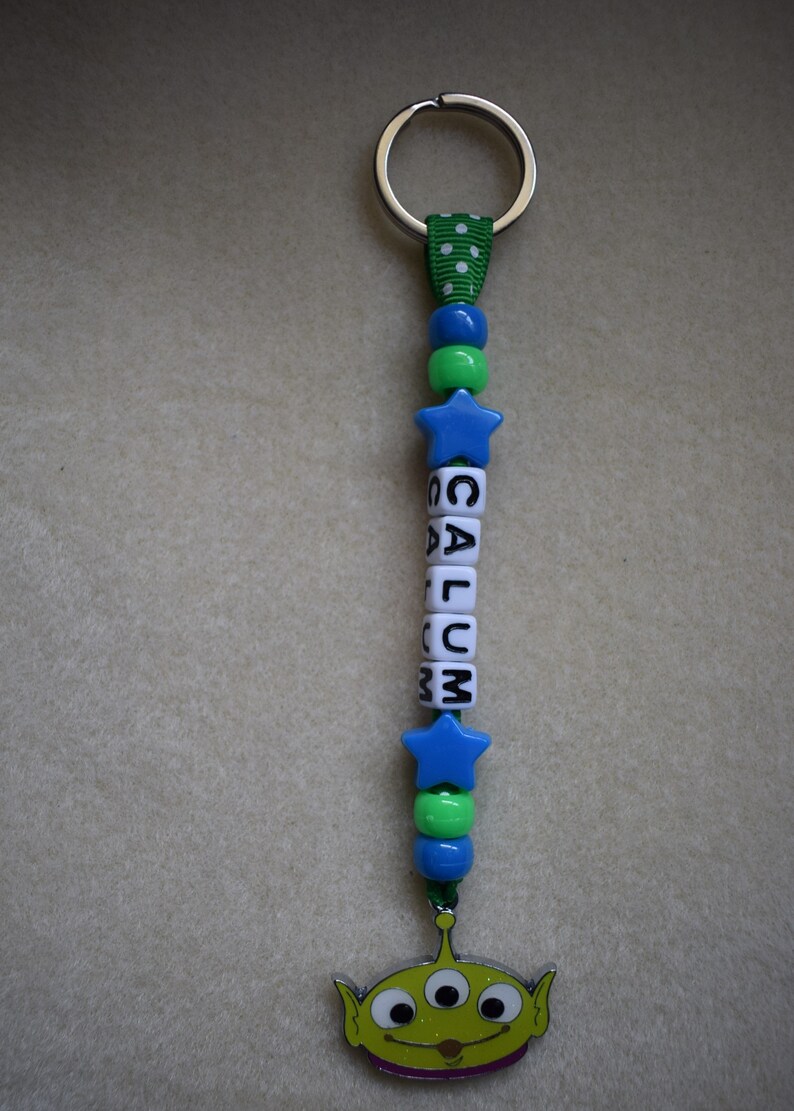 May include: A keyring featuring a green ribbon with white polka dots, blue and green plastic beads, and white cube beads spelling the name CALUM. The design includes two blue star-shaped beads and a metal charm of a three-eyed green alien character.