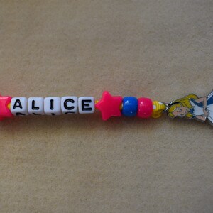 May include: A colorful beaded necklace with the name "ALICE" spelled out in white letter beads. The necklace also features a pink star bead and a charm of Alice from Alice in Wonderland.
