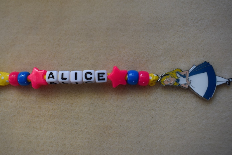 May include: A colorful beaded necklace with the name "ALICE" spelled out in white letter beads. The necklace also features a pink star bead and a charm of Alice from Alice in Wonderland.