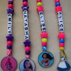 May include: Four colorful keychains with different Disney character charms. Each keychain has a name spelled out in white beads and a pink star charm. The charms are Moana, Alice in Wonderland, Tiana, and a Troll.