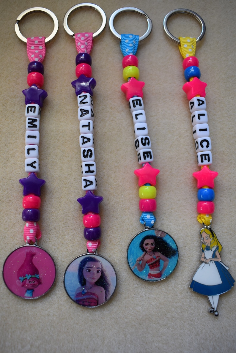 May include: Four colorful keychains with different Disney character charms. Each keychain has a name spelled out in white beads and a pink star charm. The charms are Moana, Alice in Wonderland, Tiana, and a Troll.