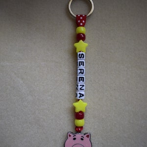 May include: A keyring featuring a silver metal ring attached to a red ribbon with white polka dots. The design includes yellow star beads, red and yellow round beads, and white cube beads spelling the name SERENA, ending with a pink pig charm.