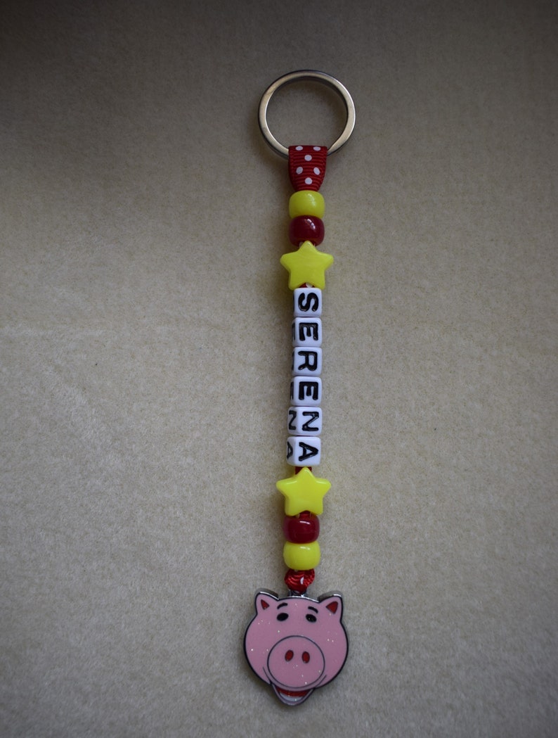 May include: A keyring featuring a silver metal ring attached to a red ribbon with white polka dots. The design includes yellow star beads, red and yellow round beads, and white cube beads spelling the name SERENA, ending with a pink pig charm.