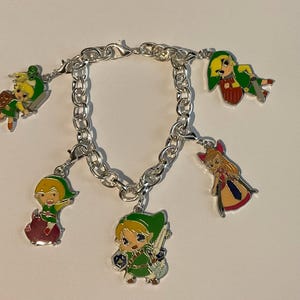 Può includere: Un bracciale con ciondoli in metallo color argento con cinque ciondoli in smalto colorato. I ciondoli raffigurano vari personaggi animati, tra cui quattro figure vestite con tuniche verdi con spade e scudi, e una figura con lunghi capelli biondi in abito.