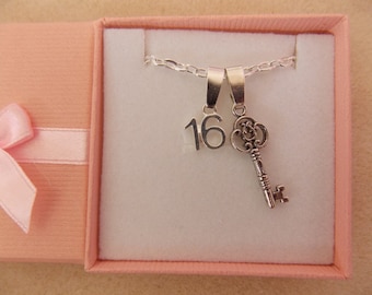 May include: A silver necklace with a key charm and a charm with the number 16 on it. The necklace is in a pink gift box with a pink ribbon.