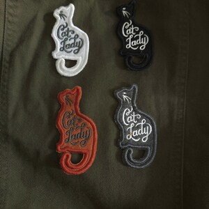 May include: Four embroidered patches shaped like sitting cats, each featuring the text Cat Lady in cursive script. The patches are white, black, orange, and gray, and are arranged in a two-by-two grid on a dark olive green fabric background.