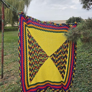 May include: A rectangular crochet blanket featuring a large central yellow triangle pattern flanked by sections of multicolored yarn in shades of green, yellow, red, and blue. The border consists of alternating stripes of purple, red, green, and blue.