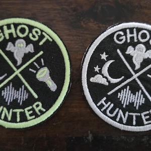 May include: Two circular embroidered patches featuring the text GHOST HUNTER. The design includes a ghost, a crescent moon with stars, a flashlight, and a sound wave icon. One patch has a light green border, while the other has a white border.