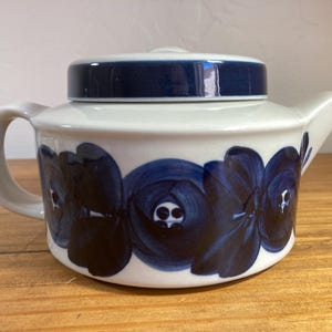 May include: A ceramic teapot with a white base and a dark blue floral pattern encircling the body. The teapot features a matching dark blue lid, a curved handle on the left side, and a short spout on the right side, all in a low-profile design.