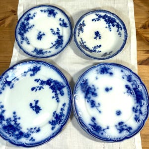 May include: Four antique white porcelain plates with a blue floral design. The plates have a scalloped edge and a detailed blue floral pattern around the rim and center. The plates are arranged on a white linen surface.