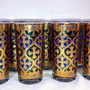 May include: A set of six cylindrical drinking glasses featuring a repeating pattern of gold filigree, deep blue, and emerald green quatrefoil shapes with central cross motifs. The glasses have a clear glass base and a gold rim at the top edge.