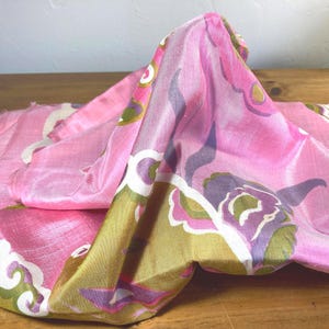 May include: A piece of fabric with a shiny, smooth texture featuring a vibrant pink base color. The textile is decorated with a repeating pattern of stylized floral shapes in shades of olive green, deep purple, and white, creating a bold visual contrast.