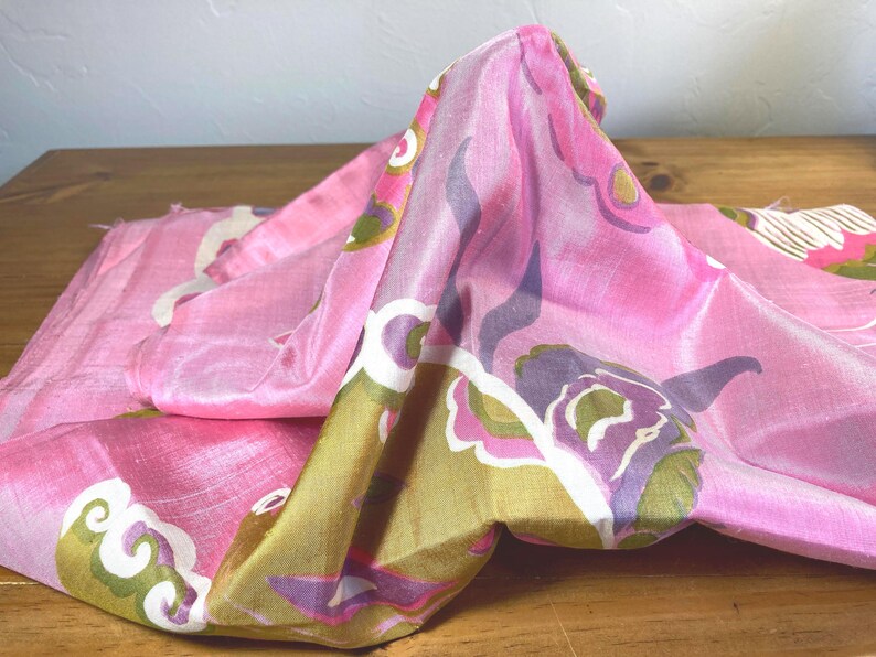 May include: A piece of fabric with a shiny, smooth texture featuring a vibrant pink base color. The textile is decorated with a repeating pattern of stylized floral shapes in shades of olive green, deep purple, and white, creating a bold visual contrast.