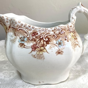 May include: A white ceramic creamer with a floral design in brown, blue, and yellow. The creamer has a curved spout and a handle. The floral pattern is around the rim and on the body of the creamer. The creamer is a vintage style.