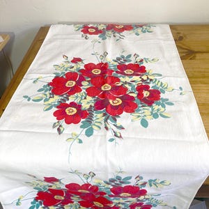 May include: A white rectangular table runner featuring a repeating floral pattern of large red blossoms with yellow centers, surrounded by green and pale yellow leaves and stems. The design is printed symmetrically across the fabric surface.