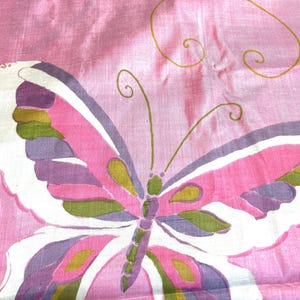 May include: A pink fabric featuring a large butterfly illustration. The butterfly has white, purple, and olive green wing segments with thin gold outlines. The fabric has a subtle sheen and a visible weave texture throughout the material.