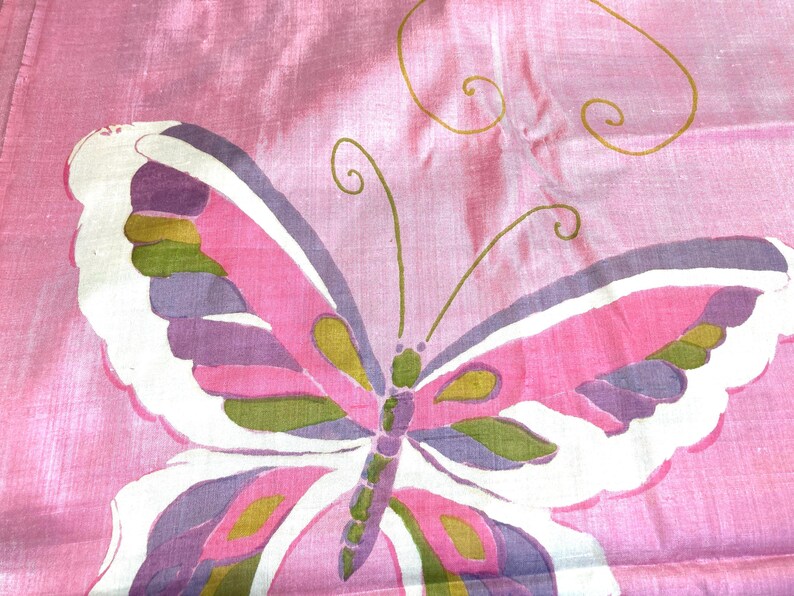 May include: A pink fabric featuring a large butterfly illustration. The butterfly has white, purple, and olive green wing segments with thin gold outlines. The fabric has a subtle sheen and a visible weave texture throughout the material.