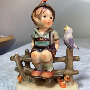 May include: A ceramic figurine of a young boy in traditional Bavarian attire, seated on a wooden fence with a small bird perched beside him. The boy wears a green shirt, brown lederhosen, and a hat with a feather. The bird is purple and white.