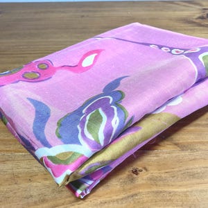 May include: A folded piece of fabric featuring a vibrant pink base color with a repeating abstract floral pattern in shades of purple, olive green, and white. The textile has a smooth, lightweight appearance with visible folds and soft edges.