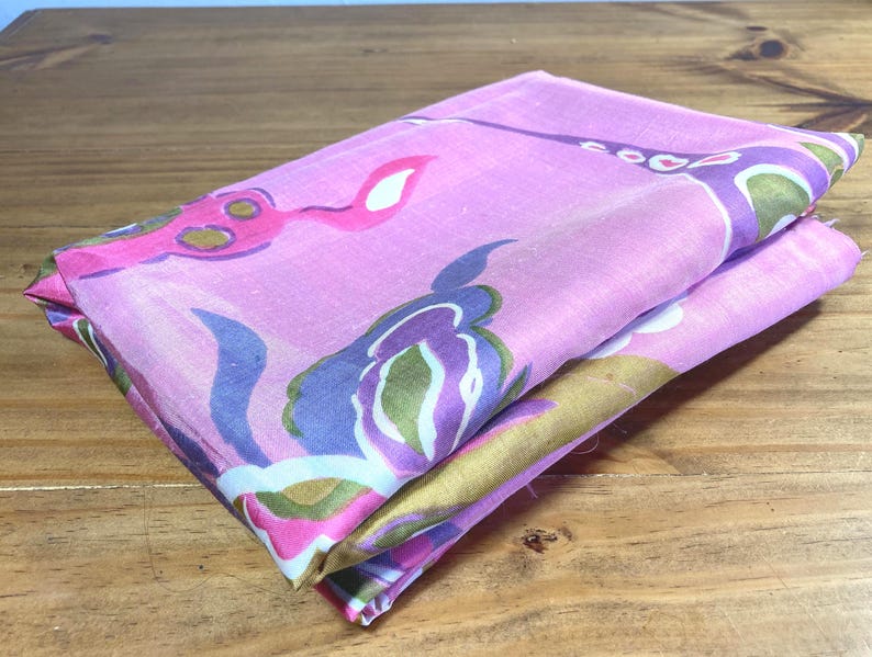 May include: A folded piece of fabric featuring a vibrant pink base color with a repeating abstract floral pattern in shades of purple, olive green, and white. The textile has a smooth, lightweight appearance with visible folds and soft edges.