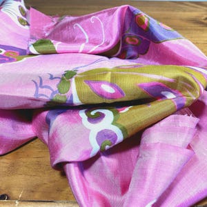 May include: A pink silk scarf featuring a repeating pattern of stylized butterflies in shades of olive green, purple, and white. The fabric has a subtle sheen and a soft, draped texture, showcasing intricate wing details and curved antenna motifs.