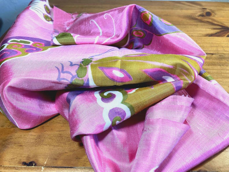 May include: A pink silk scarf featuring a repeating pattern of stylized butterflies in shades of olive green, purple, and white. The fabric has a subtle sheen and a soft, draped texture, showcasing intricate wing details and curved antenna motifs.
