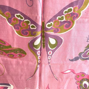 May include: A pink fabric featuring a repeating pattern of butterflies. The butterflies are rendered in shades of purple, olive green, and white with intricate swirling details. The textile has a visible weave texture throughout the surface.