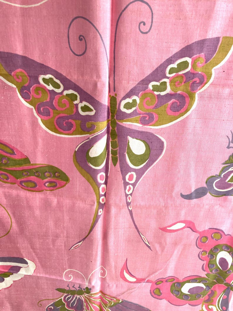 May include: A pink fabric featuring a repeating pattern of butterflies. The butterflies are rendered in shades of purple, olive green, and white with intricate swirling details. The textile has a visible weave texture throughout the surface.