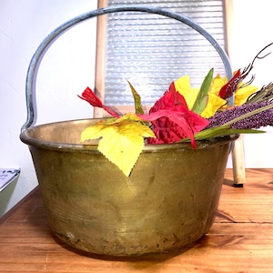 May include: A round brass bucket with a weathered patina and a sturdy metal swing handle. The bucket is filled with artificial foliage featuring vibrant red and yellow leaves along with purple textured stems, creating a colorful contrast.