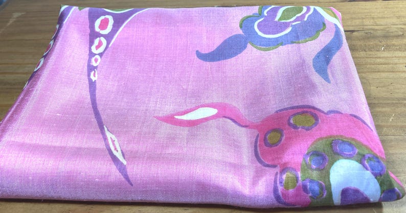 May include: A folded fabric square featuring a vibrant pink base color with a repeating pattern of abstract floral shapes in shades of purple, blue, olive green, and white. The material has a subtle sheen and a smooth, woven texture throughout.